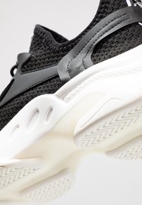 Zapatilla deportiva de malla negra con parte superior texturizada, detalles en gris contrastante y una suela blanca flexible con dibujo de patrón.