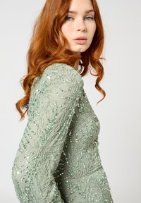 Groene versierde jurk met ingewikkelde bladderspatronen, pailletten en lange mouwen. De jurk heeft een getailleerde pasvorm en gestructureerde stof.