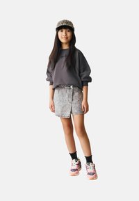 Bambino con cappellino a stampa leopardata, felpa grigia oversize, gonna in denim acid-wash, calzini neri e sneaker rosa a stampa leopardata, in piedi contro uno sfondo bianco.
