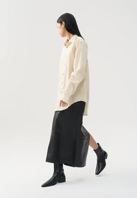 Beige Hemdbluse mit lockerer Passform, kombiniert mit einem schwarzen Leder-Midi-Rock und spitzen schwarzen Ankle-Boots, die eine strukturierte Silhouette schaffen.