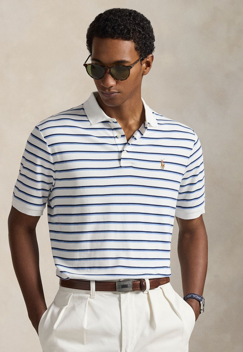 Polo blanc à rayures bleues, manches courtes, col à deux boutons. Porté avec un pantalon beige et une ceinture marron ; des lunettes de soleil viennent compléter le look.