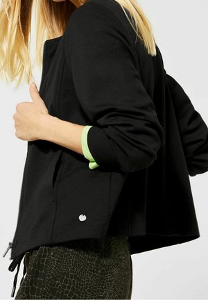 Femme aux cheveux blonds portant une veste noire zippée par-dessus un chemisier vert à manches longues et un pantalon vert foncé texturé, vue de profil.