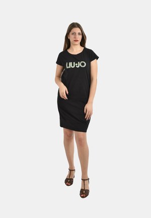 Donna in piedi che indossa un vestito nero a maniche corte con il logo "LIU·JO" e sandali marroni aperti in punta con cinturino a T su sfondo bianco.