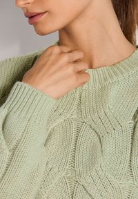 Frau trägt einen hellgrünen, grobstrickigen Pullover und berührt mit der rechten Hand den Halsausschnitt in der Nähe von Hals und Kinn.