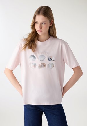 Giovane donna con capelli lunghi indossa una T-shirt rosa chiaro con sei illustrazioni di conchiglie marine e la parola "Ocean", abbinata a jeans scuri.