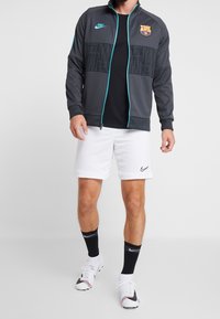 Szara kurtka Nike z zamkiem błyskawicznym i akcentami w kolorze teal, z logo FC Barcelona, w zestawie z czarną koszulką, białymi szortami i czarnymi skarpetkami.