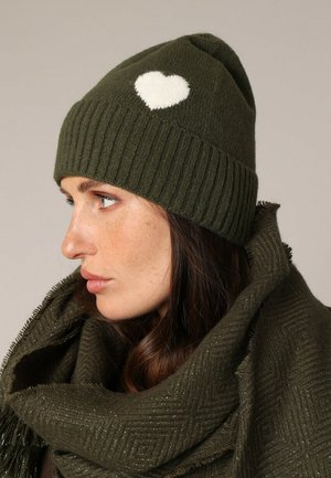 Bonnet en tricot vert olive avec une texture côtelée et un accent en forme de cœur blanc. Porté avec une écharpe texturée verte assortie.