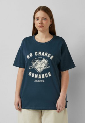 Jeune femme aux longs cheveux bruns portant des boucles d'oreilles argentées en forme de cercle et un t-shirt bleu marine avec un motif vintage de cœur et texte romantique.