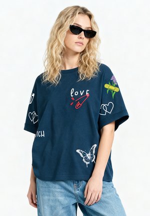 Blonde vrouw met golvend haar die zwarte zonnebril draagt, een donkerblauw T-shirt met liefde, vlinder-, hart- en bloempatches, en blauwe spijkerbroek.