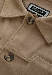 Giacca in suede beige con colletto, chiusura a bottoni e tasca sul petto. Presenta una consistenza morbida e dettagli di cucitura prominenti. Etichetta visibile.