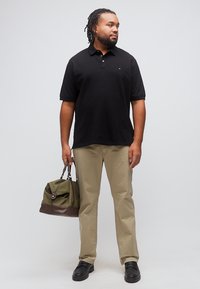 Άνδρας με πλεξούδες, φορώντας μαύρο πόλο πουκάμισο, χακί παντελόνι, μαύρα παπούτσια, κρατώντας πράσινη και καφέ τσάντα duffel, στέκεται μπροστά σε απλό φόντο.