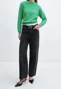 Groene gebreide trui met textuur, gecombineerd met hoge zwarte denimjeans en zwarte puntige hakken. De outfit bevat een riem met een zilveren gesp.