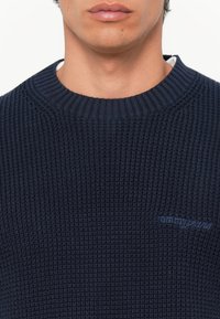 Donker marineblauwe gebreide trui met ribbeltextuur, ronde hals, en geborduurd "tommyjeans" logo in het blauw op de linkerborst.