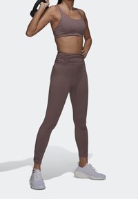 Brun træningscrop top og høj midje leggings med en ruché design. Modellen holder en kettlebell; hvide træningssko fuldender outfittet.