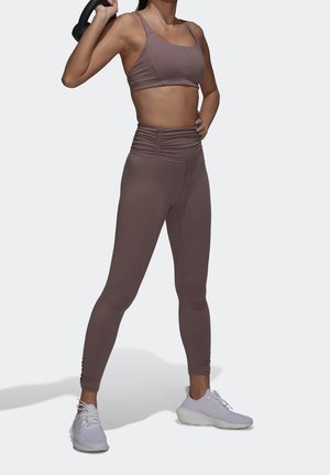 Brun træningscrop top og høj midje leggings med en ruché design. Modellen holder en kettlebell; hvide træningssko fuldender outfittet.