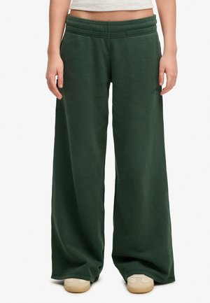 Personne portant un pantalon de survêtement large vert foncé et des baskets beiges, debout, les bras détendus le long du corps.