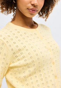 Γυναίκα με σγουρά μαλλιά φοράει κίτρινο κουμπωτό ζακέτα με μικρό μοτίβο με άνθη και χρυσό σκουλαρίκι κρίκο, κοιτάζοντας ελαφρώς προς τα αριστερά.