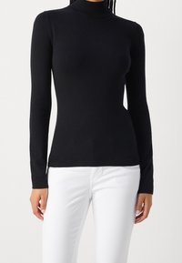 Svart långärmad turtlenecksweater i mjukt tyg med en figurnära design, med ribbad halskrage och muddar. Bärs med vita byxor.