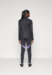 Svart langermet topp og tettsittende svarte bukser med lilla og grønne sideaccenter. Glatt stoff, atletisk design og buede sømmer.