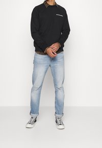 Sudadera negra de cuello redondo con logo blanco, llevada sobre una camisa de cuadros marrón, combinada con jeans de color azul claro y zapatillas en blanco y negro.
