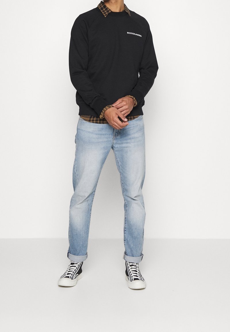 Sudadera negra de cuello redondo con logo blanco, llevada sobre una camisa de cuadros marrón, combinada con jeans de color azul claro y zapatillas en blanco y negro.