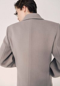 Grå uldfrakke med en struktureret silhuet, brede revers og puffede ærmer, der har en glat tekstur og rene syninger.