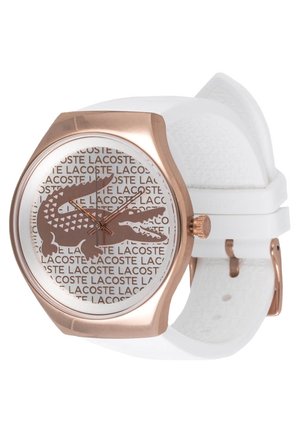 Montre ronde avec boîtier en or rose, bracelet blanc et cadran avec un grand logo crocodile marron sur le texte répété "LACOSTE".