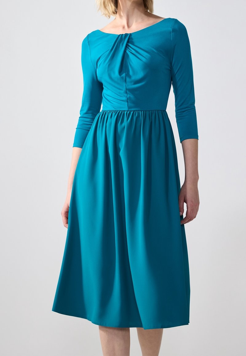Robe bleu sarcelle longueur genou avec un col rond, des manches trois-quarts, une jupe froncée et un détail de tissu torsadé sur le corsage. Texture lisse.