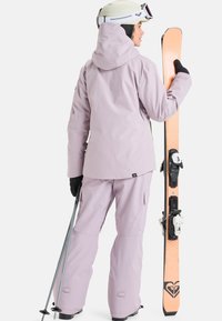 La tenue de ski comprend une veste et un pantalon imperméables de couleur violet clair, associés à des gants noirs. Un bâton de ski et des skis orange sont tenus dans la main droite.