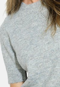 Pull en tricot gris avec un col rond et des manches courtes. La texture est douce avec un motif légèrement chiné et des fils métalliques discrets.