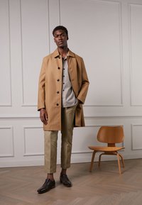 Trench beige con botones negros, camisa gris debajo, pantalones beige y zapatos oscuros; texturas suaves contra un fondo minimalista.