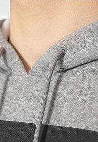 Felpa grigia con cappuccio e cordino, caratterizzata da una superficie liscia e una striscia nera orizzontale. Vista ravvicinata del collo e dei dettagli del tessuto.