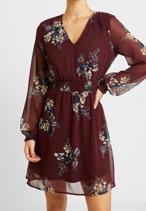 Frau trägt ein bordeauxrotes Blumenkleid mit langen transparenten Ärmeln, elastischem Taillenband und V-Ausschnitt, Hand auf der Hüfte ruhend.