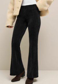 Schwarze Cordflügelhose mit vertikalen Nähten, hochgeschnittenem Design und weicher Textur, kombiniert mit einer cremefarbenen Kunstfelljacke.