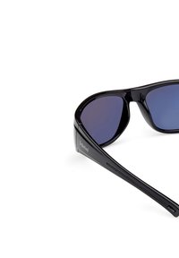 Gafas de sol negras con lentes azules en degradado, montura de plástico elegante y un logo de Timberland en el lateral. Diseño curvado con textura suave.