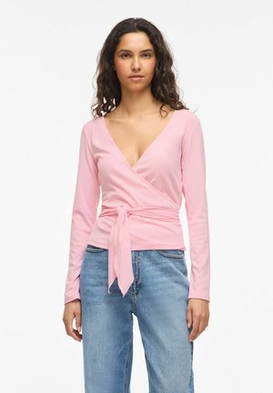 Femme aux cheveux bouclés portant un top croisé rose clair noué à la taille et un jean bleu, debout devant un fond blanc.