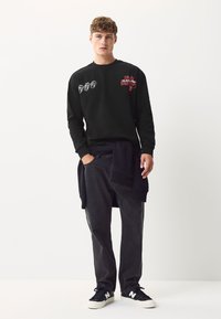 Sort langærmet skjorte med grafisk tryk i rød og hvid; kombineret med mørkegrå bukser og sorte sneakers med hvidt logo.