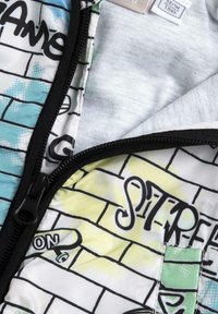 Chaqueta de cremallera colorida con impresión de estilo graffiti sobre un fondo de ladrillos blancos, que presenta tonos de azul, verde y acentos amarillos.