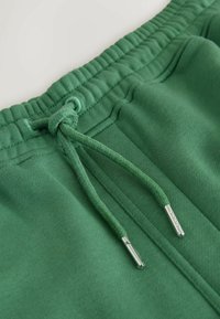 Taille de pantalon de survêtement verte avec cordon noué et embouts métalliques, montrant un tissu texturé et des détails de couture.