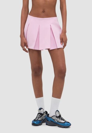 Person, die einen kurzen rosa plissierten Rock, weiße Kniestrümpfe und blaue und graue New Balance Sneaker vor einem schlichten Hintergrund trägt.