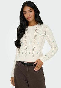 Pull en maille de couleur crème avec un motif zigzag et des broderies florales bleues. Doté de manches longues et d'un col rond.