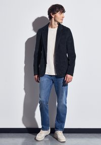 Black corduroy blazer with notched lapels over a white t-shirt, paired with light blue jeans and white sneakers. Smooth texture, tailored fit.