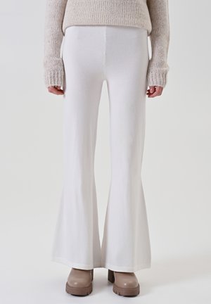 Person trägt hochgeschnittene weiße Schlaghosen, beige klobige Stiefel mit Absatz und einen beigen gestrickten Pullover mit langen Ärmeln.