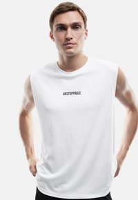 Młody mężczyzna w bezrękawniku w kolorze białym z napisem "UNSTOPPABLE", pozujący z rękami na biodrach na jednolitym tle.