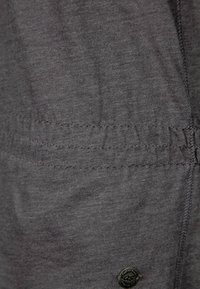 Tissu gris à texture lisse, avec un motif cousu et une petite étiquette ronde noire avec logo. Vue en gros plan, mettant en valeur les détails.