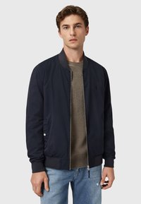 Chaqueta bomber de la Armada con puños y dobladillo acanalados, cierre con cremallera, bolsillos laterales y un logo sutil en el pecho; usada sobre un suéter gris.