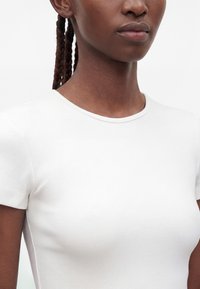 Nahaufnahme einer Person, die ein schlichtes weißes, geripptes Kurzarm-T-Shirt trägt, mit geflochtenem Haar und neutralem Gesichtsausdruck.