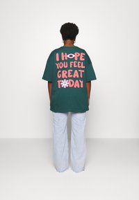 Groene oversized t-shirt met grote roze tekst op de achterkant met de tekst "IK HOOP DAT JE JE HEEL GOED VOELT VANDAAG" en een bloemenafbeelding. Draagt met lichte jeans.