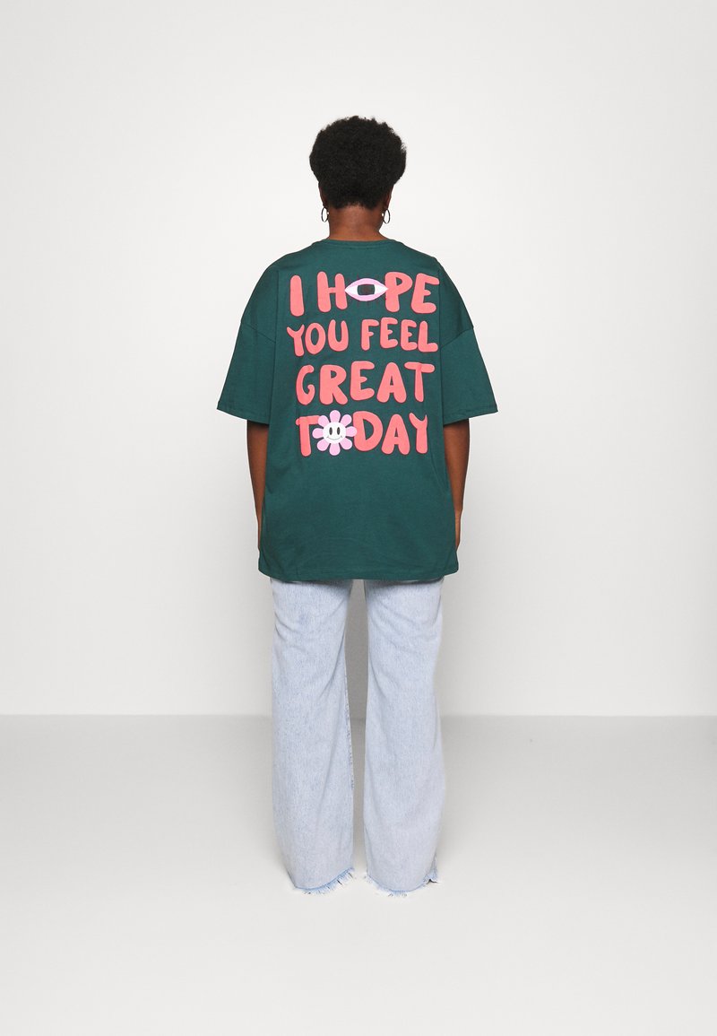 Groene oversized t-shirt met grote roze tekst op de achterkant met de tekst "IK HOOP DAT JE JE HEEL GOED VOELT VANDAAG" en een bloemenafbeelding. Draagt met lichte jeans.
