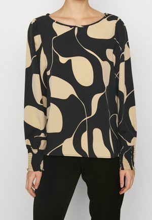 Vrouw draagt een blouse met lange mouwen met een beige en zwarte abstracte print en zwarte broek, getoond van nek tot heupen.